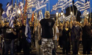 Man in camo trousers stands to attention in front of sea of Greek flag-wavers